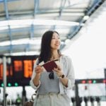 A young woman in an airport terminal holding a passport and travel documents, symbolizing the importance of choosing the right coverage, such as travel insurance vs health insurance, for international trips.