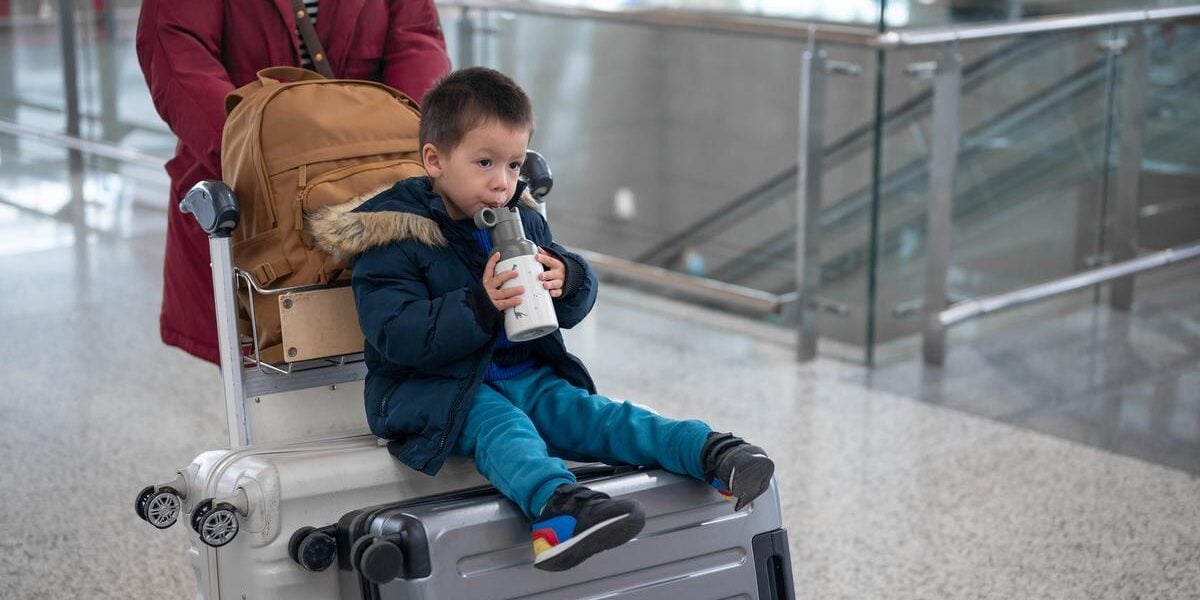 a toddler drinking from a reusable water bottle while sitting on a suitcase trolley, one of many great holiday travel tips