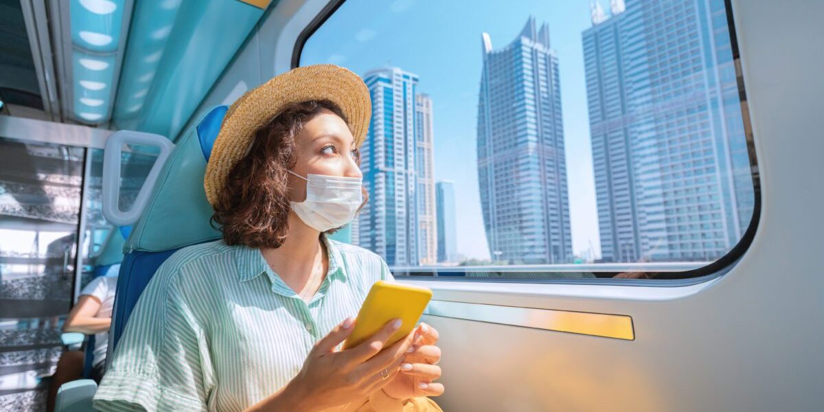 Woman in a protective medical mask rides the subway, holds a smartphone in her hands and looks out the window where the giant skyscrapers of Dubai rise