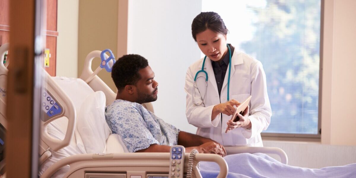 a male patient laying in a hospital bed talking to a female doctor, reflecting on the importance of annual health insurance reviews for ensuring adequate hospital coverage