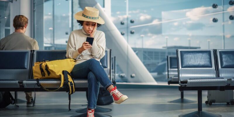 A stressed woman sitting on a chair in the airport, hand on her face in shock, highlighting the need for trip cancellation insurance.