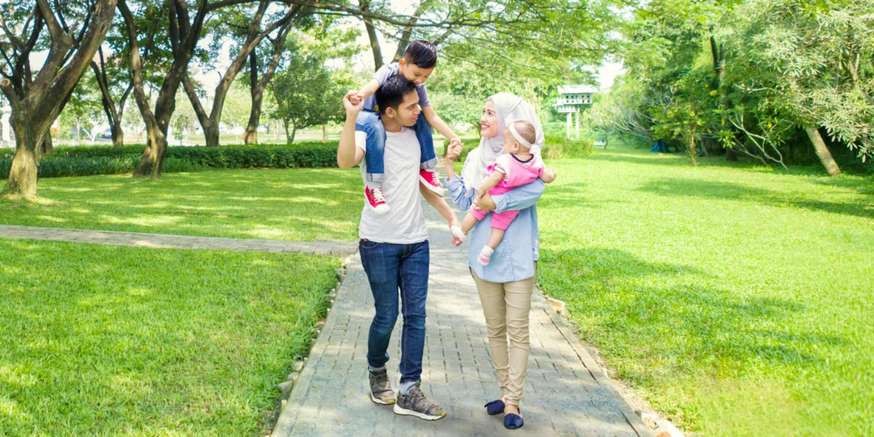 a happy Indonesian couple walking along a footpath in a lush green park carrying their two small children