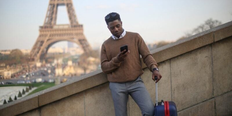 a Bahamian man standing in front of the Eiffel Tower with a suitcase, looking at his phone to check the wording of his insurance for Bahamian citizens