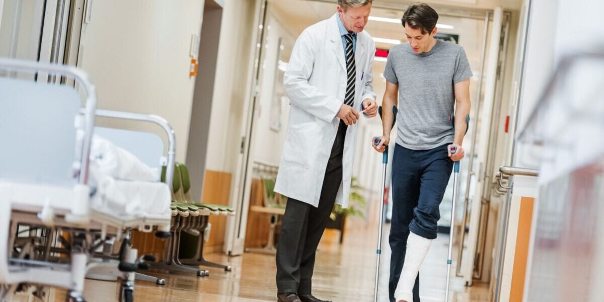 a patient walking on crutches alongside a doctor in a country that offers universal healthcare